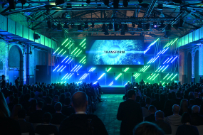 Publikum in dunkler Business-Kleidung sieht in einem Saal zu einer Bühne auf, wo ein Banner mit dem Wort "TRANSFORM" und einem sternähnlichen visuellen Effekt präsentiert wird. Die Szene ist in blaues und grünes Licht getaucht.